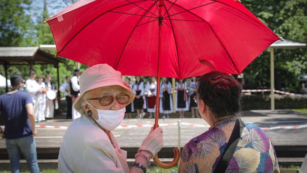 Две женщины, одна из которых носит лицевую маску для борьбы с распространением коронавируса, защищают себя от солнца под зонтом, наблюдая, как фольклорная группа исполняет танец летнего солнцестояния в музее деревни Дмитрия Густи в Бухаресте, Румыния - Sputnik Moldova-România