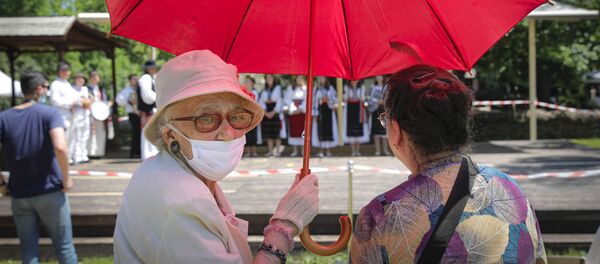Две женщины, одна из которых носит лицевую маску для борьбы с распространением коронавируса, защищают себя от солнца под зонтом, наблюдая, как фольклорная группа исполняет танец летнего солнцестояния в музее деревни Дмитрия Густи в Бухаресте, Румыния - Sputnik Moldova-România