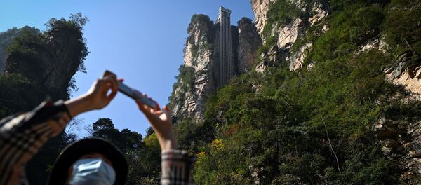 A tourist with a face mask takes a picture with her mobile phone at the entrance of the Bailong Elevator in the Zhangjiajie National Forest Park in China's Hunan province. - Sputnik Moldova