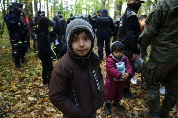 Un copil migrant irakian stă în picioare în timp ce este înconjurat de polițiști de frontieră și ofițeri de poliție după ce au trecut frontiera bielorusă-poloneză în timpul crizei imigranților în curs, în Hajnowka, Polonia, 14 octombrie 2021. - Sputnik Moldova-România